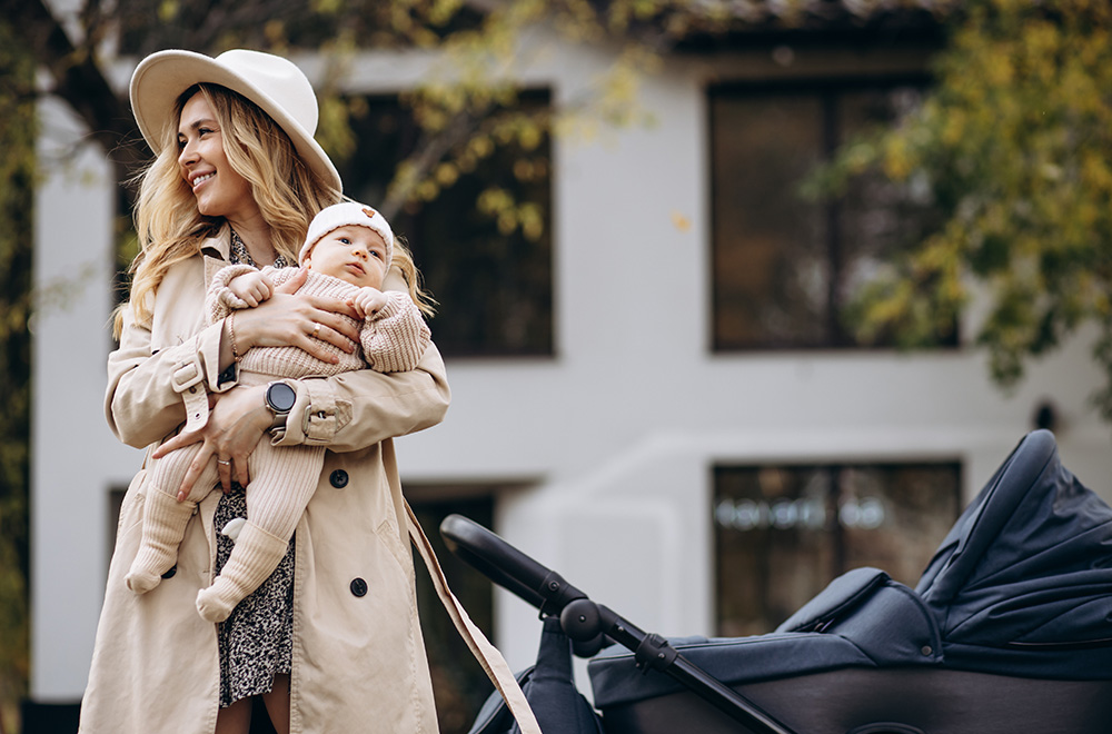 Sieviete tur bērnu rokās un iet garām ratiņiem pie ēkas.
