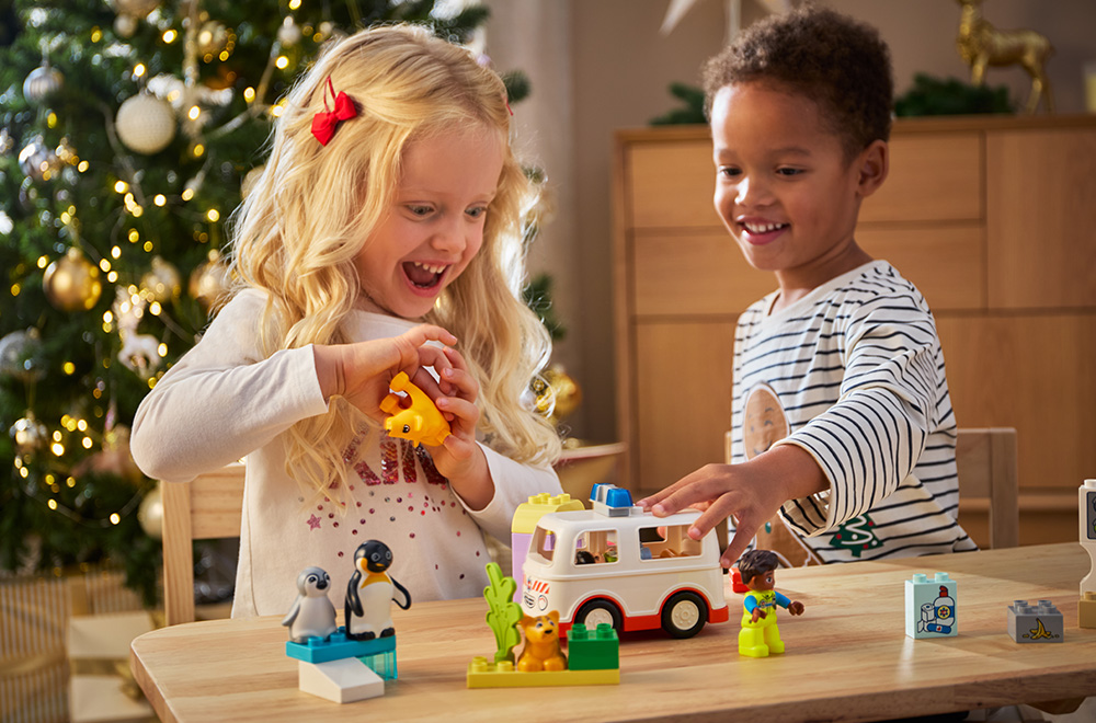 Divi bērni spēlējas ar LEGO klucīšiem zem Ziemassvētku eglītes.